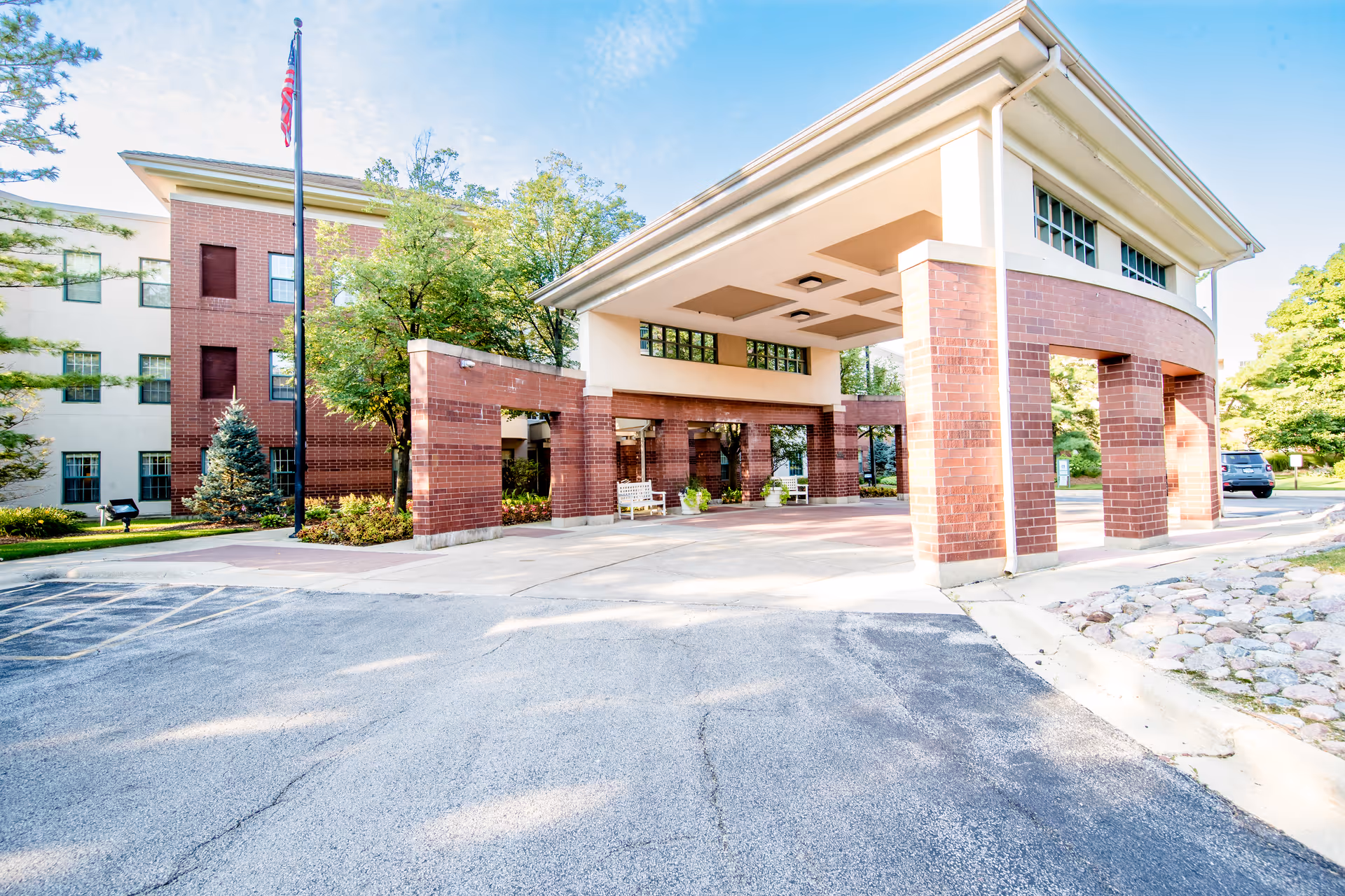 Entrance of a senior living facility named Wyndemere featuring a covered drop-off area with brick pillars, an American flag on a flagpole, surrounding trees, and a paved driveway.