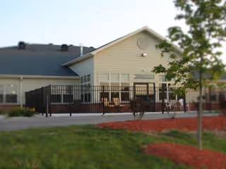 Exterior view of a single-story building with beige siding and a dark roof, featuring a fenced patio area with outdoor seating. There is a tree and landscaped grass with red mulch in the foreground.
