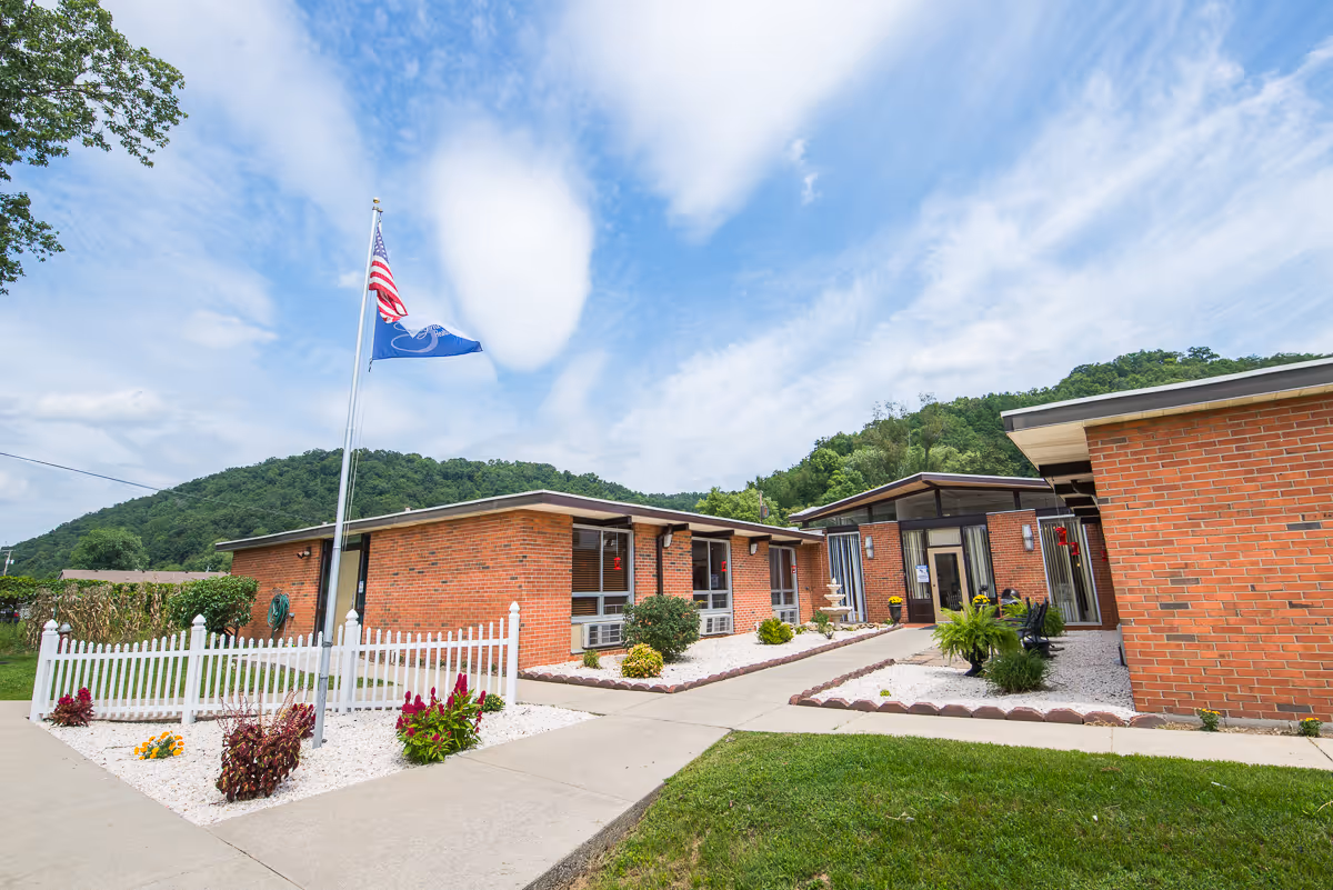 Exterior view of Prestonsburg Health Care Center showing a single-story brick building with a white picket fence, landscaped garden beds with flowers, a flagpole with the American flag and another flag, and a clear blue sky with some clouds and green hills in the background.