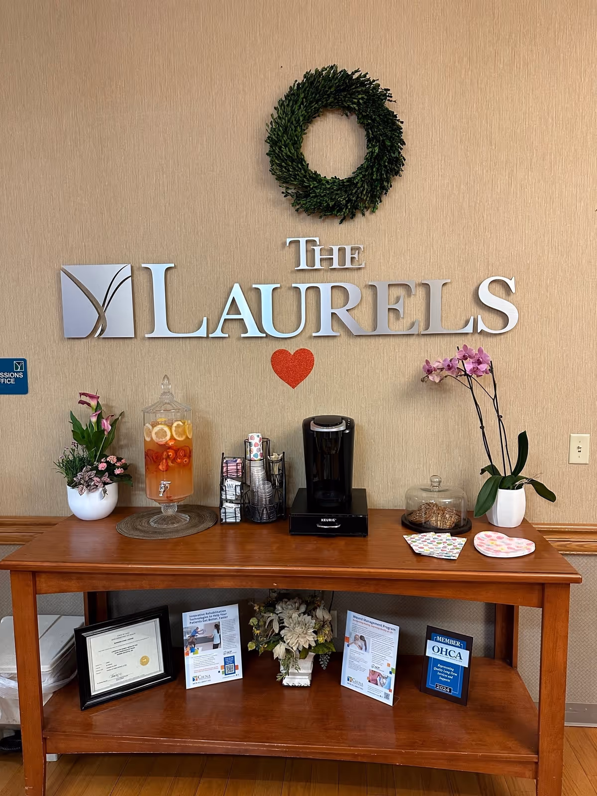 A wooden table against a beige wall with a green wreath and silver letters spelling 'THE LAURELS' with a red heart underneath. On the table are a beverage dispenser with fruit-infused water, a Keurig coffee maker, cups, napkins, a glass container with cookies, and two potted plants with flowers. Below the table are framed certificates, informational brochures, and a flower arrangement.