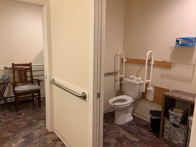 Accessible restroom with a toilet, white support grab bars, shelving, and a chair visible in the adjoining room.