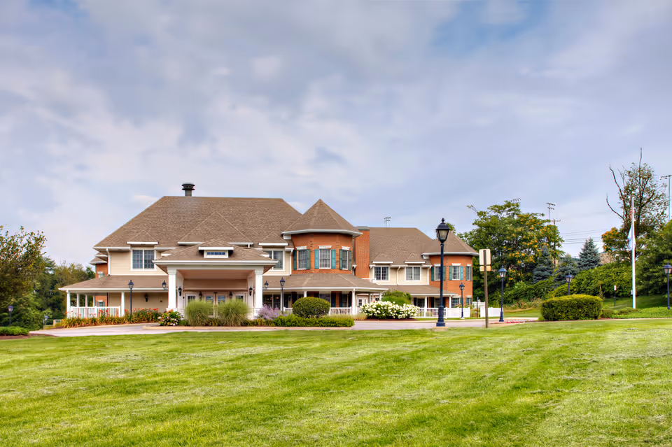 Exterior view of Juniper Village at Monroeville, a large two-story building with a combination of brick and beige siding, surrounded by well-maintained green lawns and landscaping under a cloudy sky.