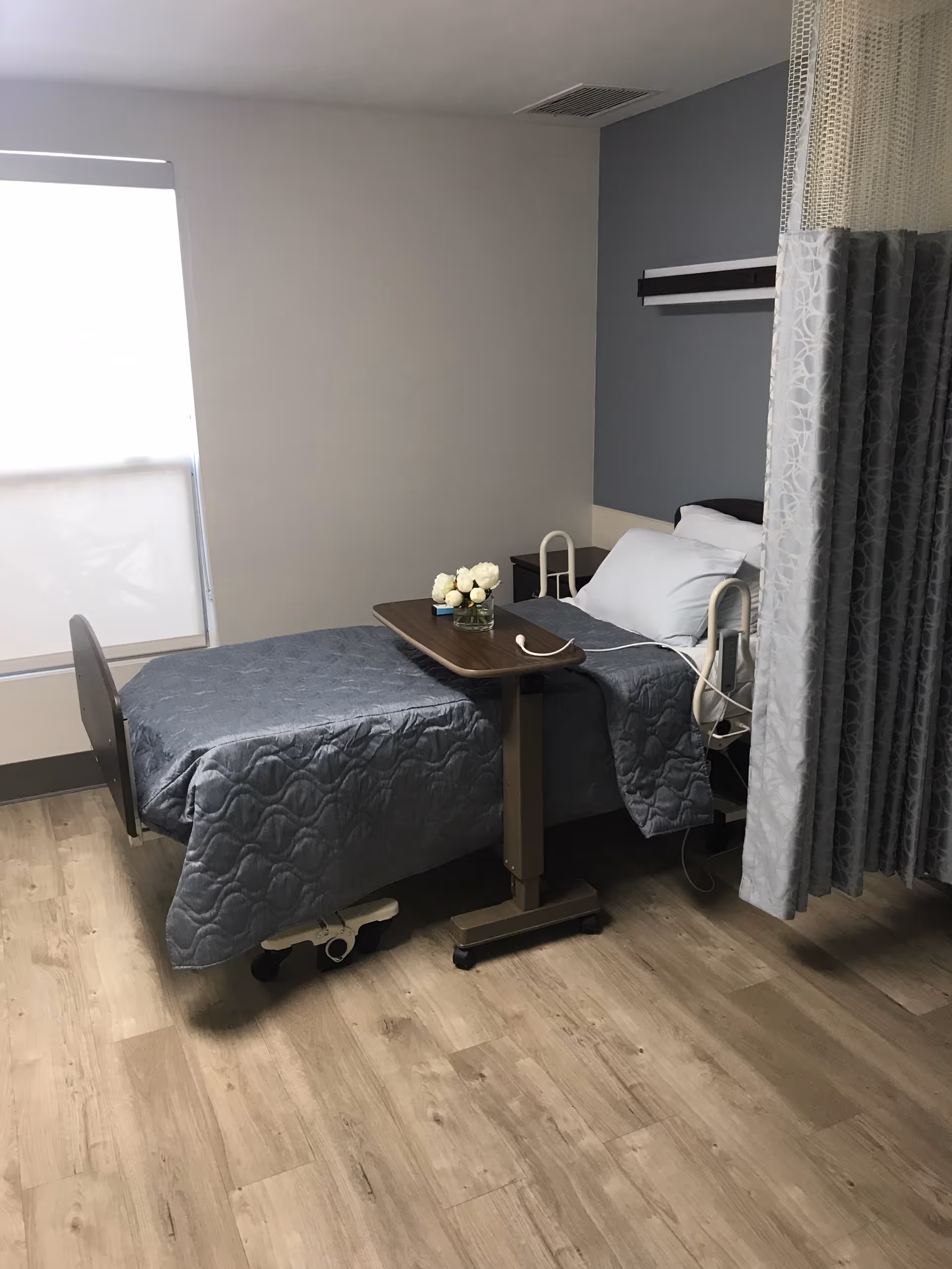 Patient room with a single hospital bed covered in a blue quilt, an overbed table with flowers, a privacy curtain and a window.