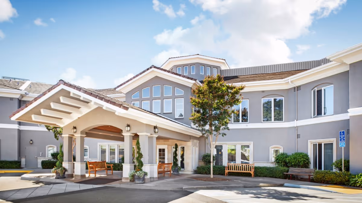 Exterior view of a senior living facility named Ivy Park at Santa Rosa, showing a covered entrance with benches, a tree, and a two-story building with multiple windows under a partly cloudy sky.