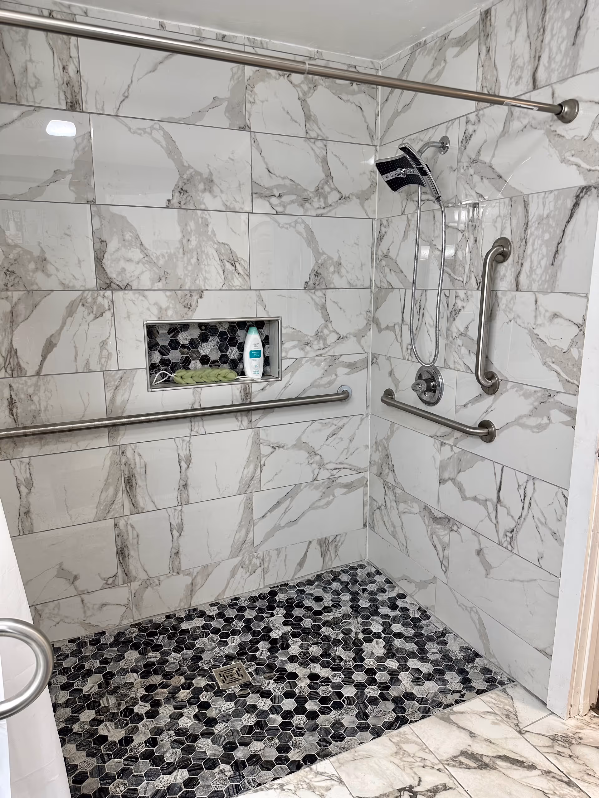 Accessible marble-tiled bathroom shower with grab bars, a niche, and a hexagonal mosaic floor.
