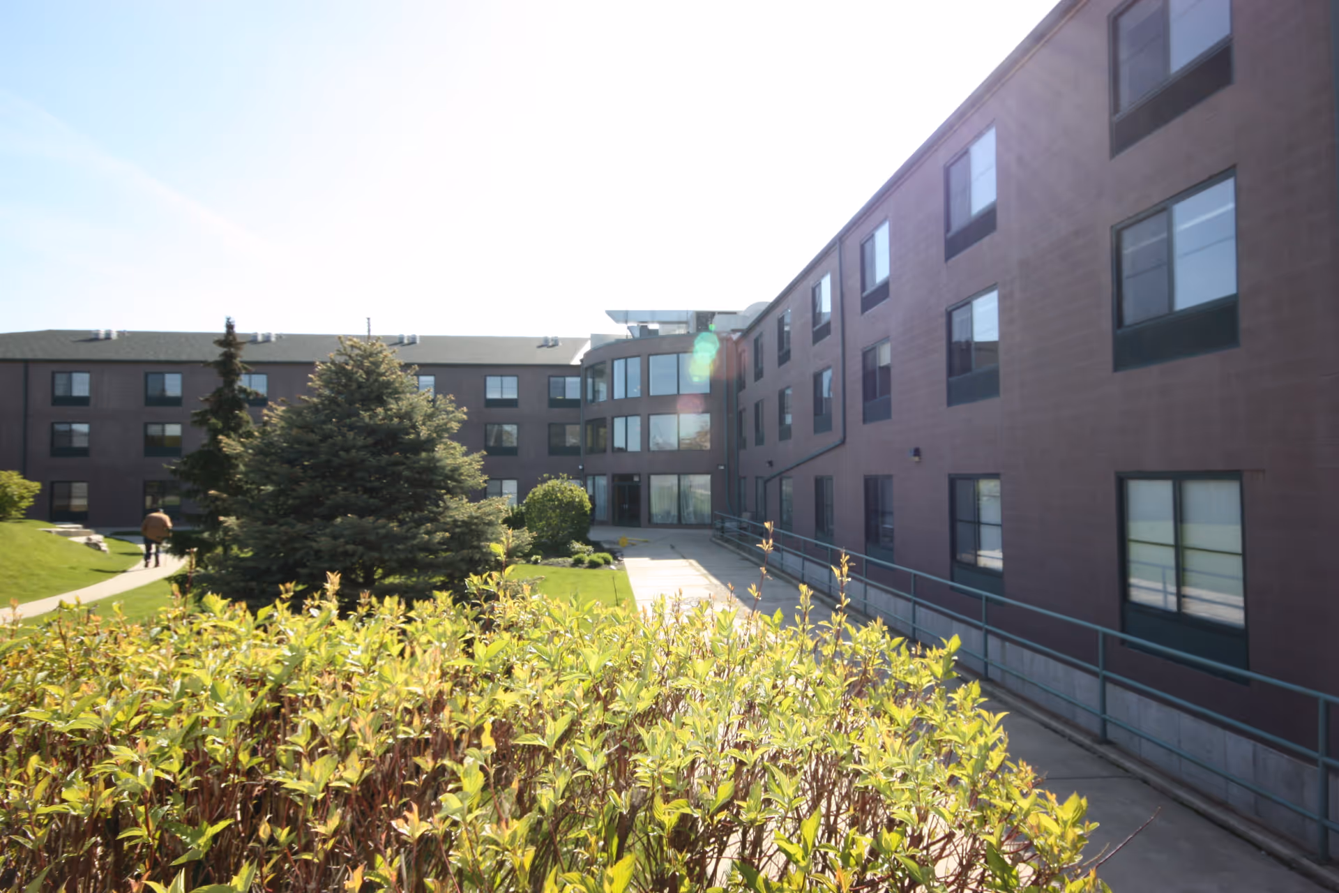 Three-story brick senior living building with a central courtyard, shrubs, a walkway, and a ramp entrance.