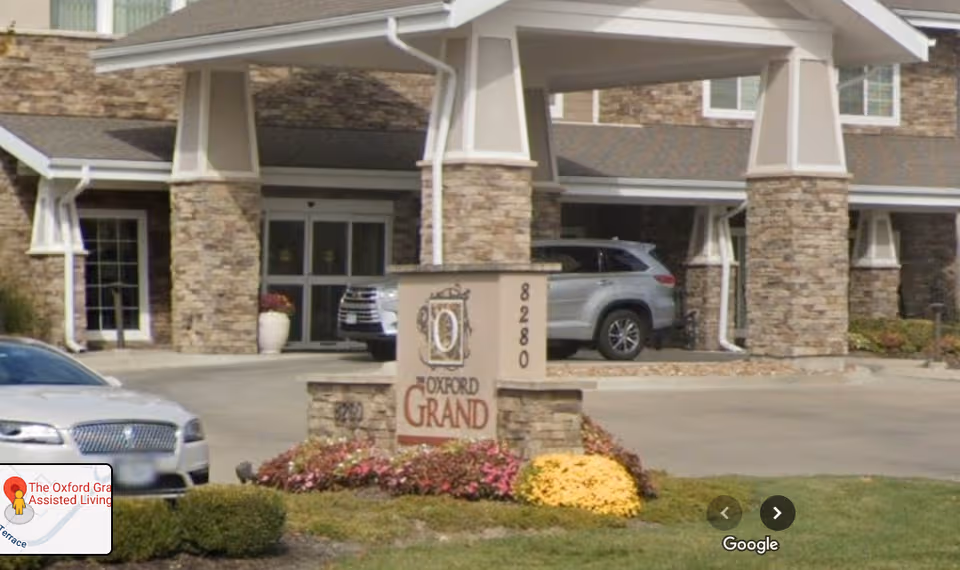 Entrance to The Oxford Grand senior living with stone facade and parked cars.