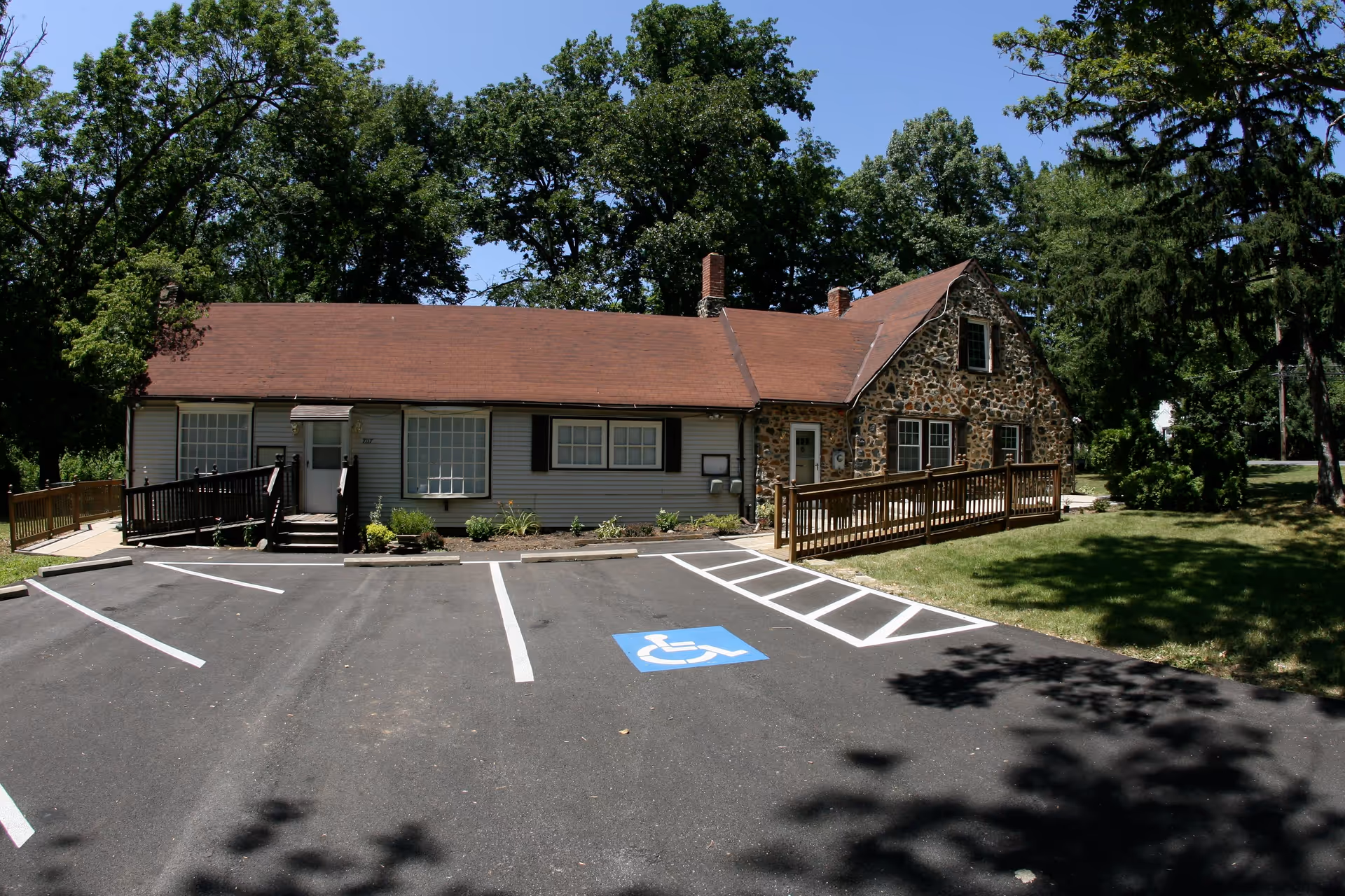 Front view of a single-story stone-and-siding building with a wooden ramp and an accessible parking space, set among trees.