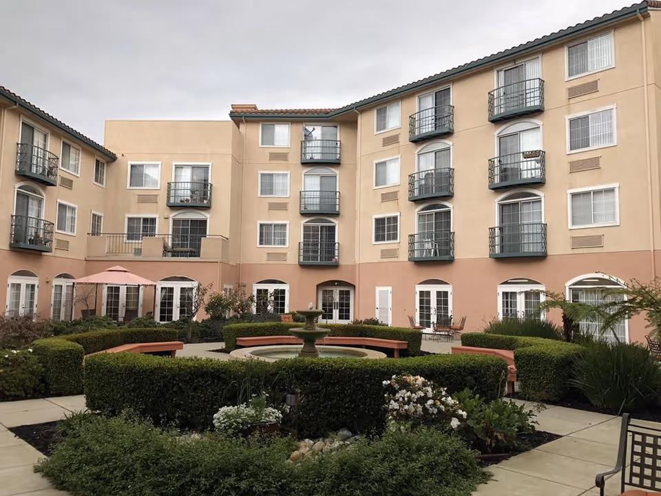 Exterior courtyard of a senior living building with balconies, hedges, and a central fountain.