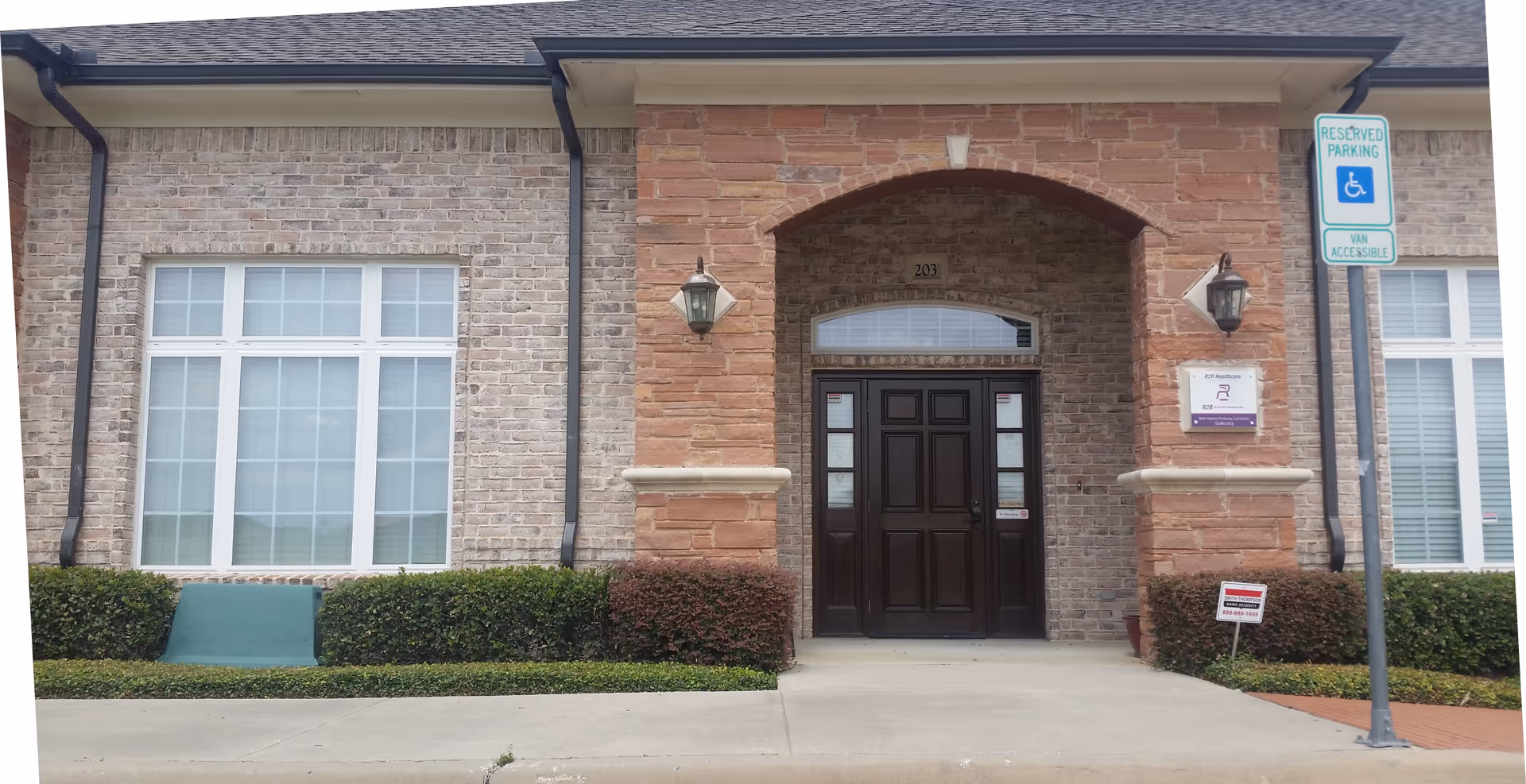 Facility entrance with brick archway, dark wooden doors, shrubs, and a handicap parking sign.