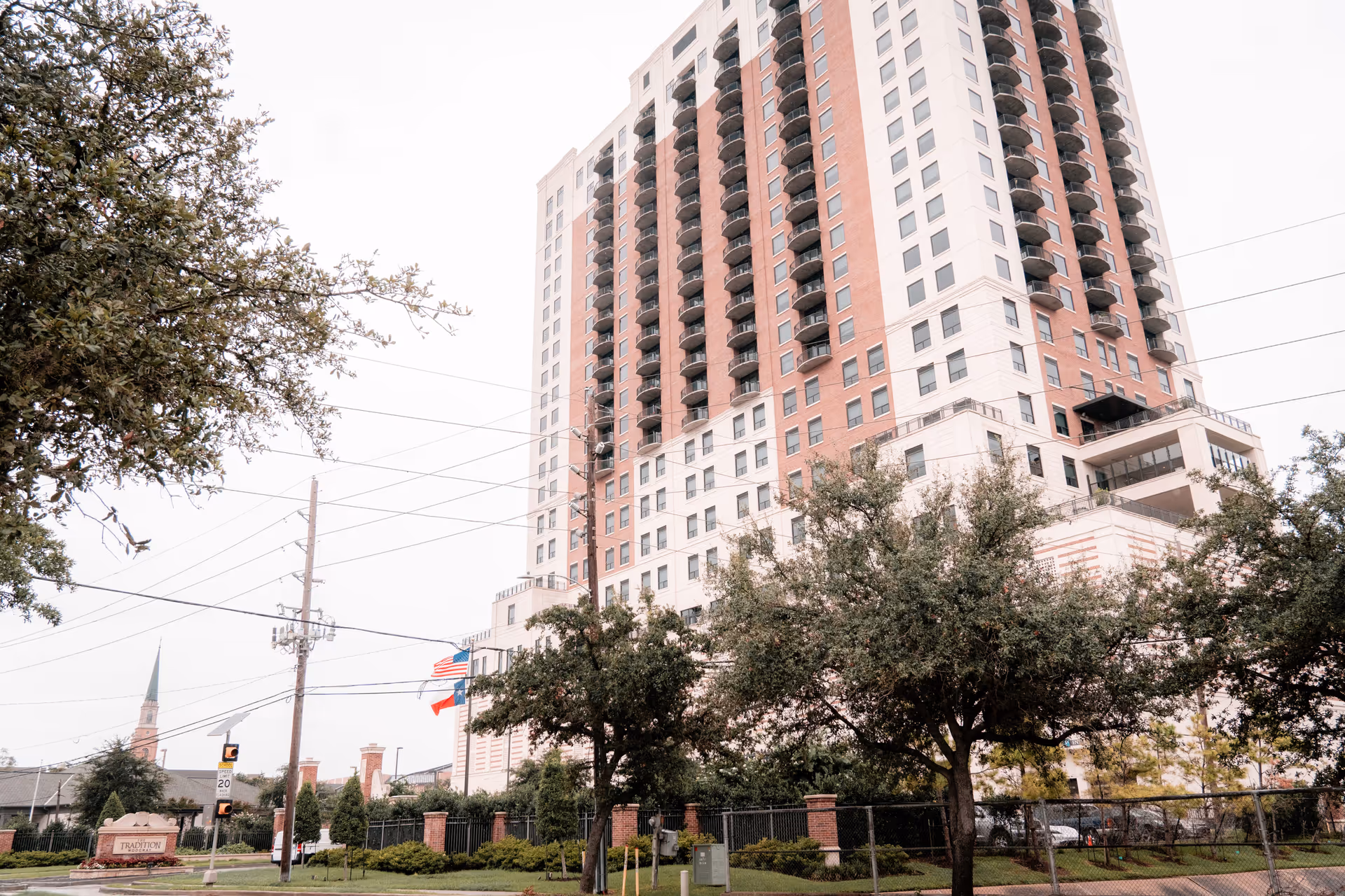High-rise residential building with balconies seen from the street, framed by trees and flags in the foreground.