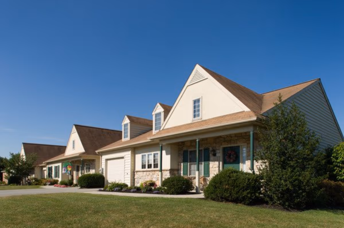 Exterior view of a residential-style building with beige siding, stone accents, green shutters, and a brown roof under a clear blue sky. The building has a porch with green pillars and is surrounded by well-maintained bushes and grass.