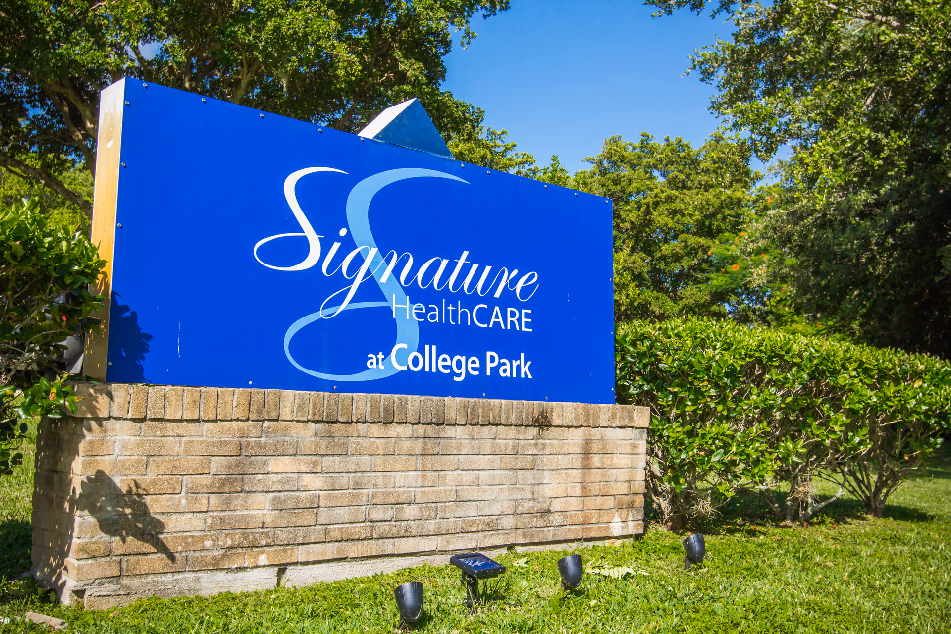 Blue Signature HealthCARE sign on a brick pedestal among trees and hedges.