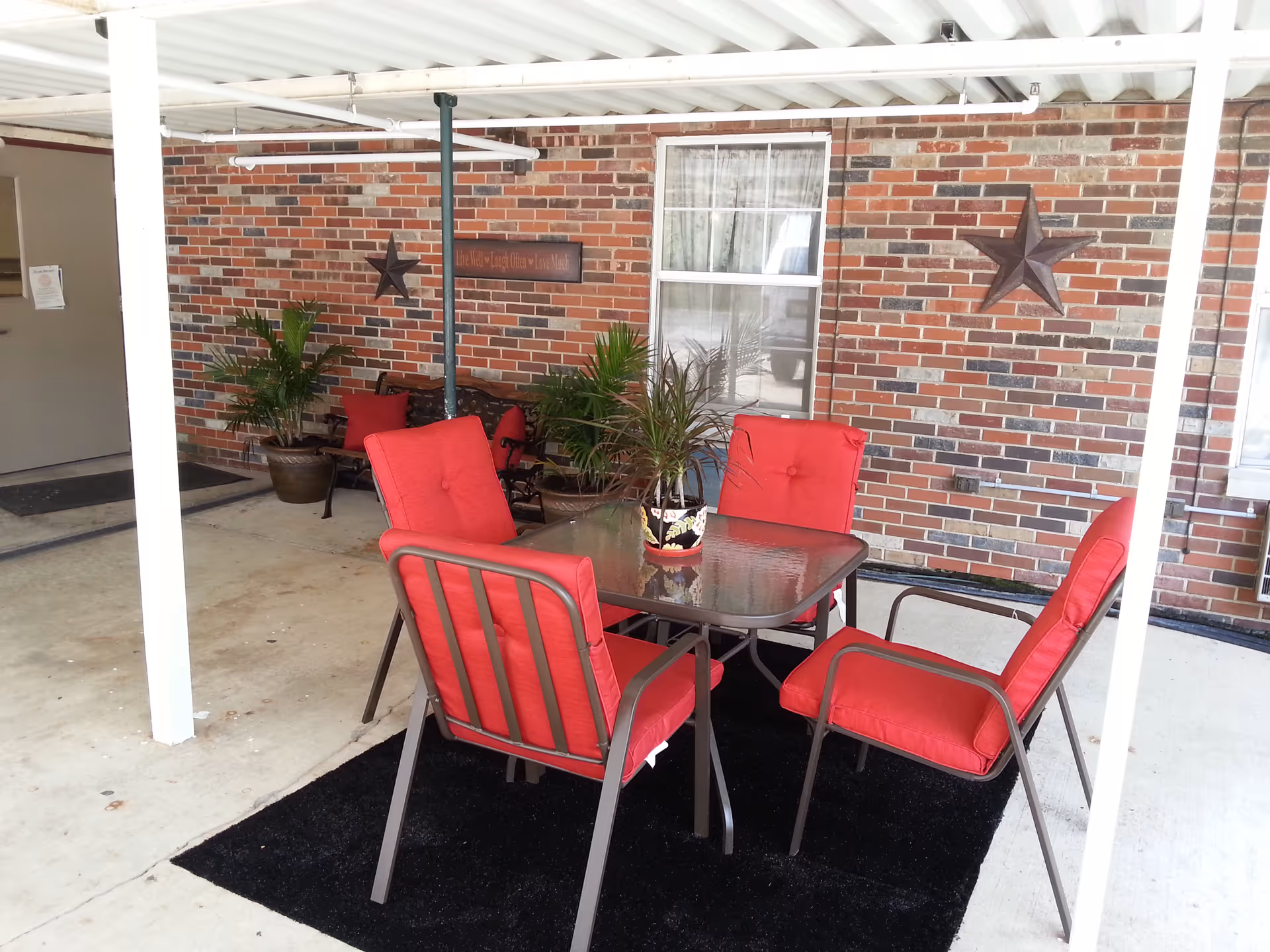 Covered outdoor patio area with a glass-top table surrounded by four red cushioned chairs. There are potted plants on the table and in the background, against a brick wall decorated with two metal star ornaments and a wooden sign. The floor is concrete with a black rug under the table and chairs.