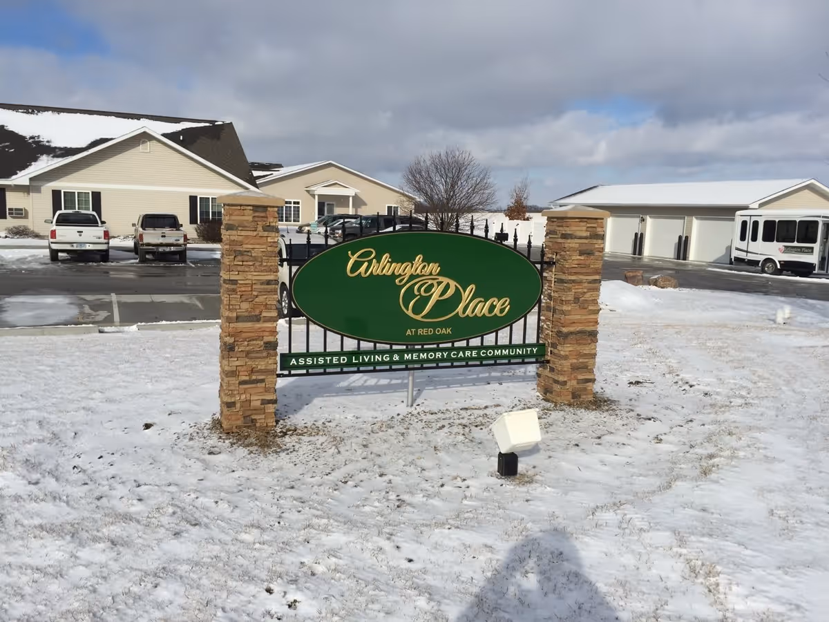 Outdoor view of Arlington Place at Red Oak assisted living and memory care community sign with snow-covered ground, parked vehicles, and buildings in the background under a cloudy sky.