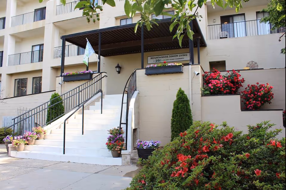 Front entrance of a senior living building with stairs, railings, and blooming planters.