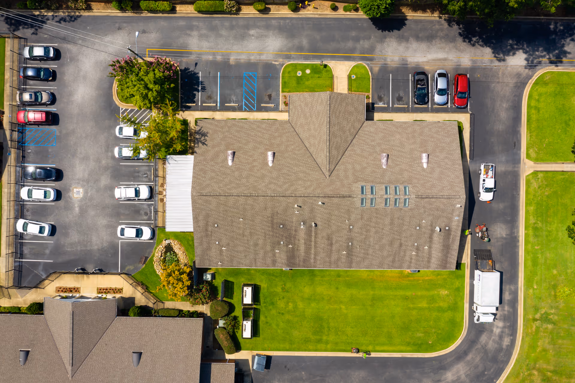 Aerial view of a senior living facility showing the building's roof, surrounding parking lots with several parked cars, green lawns, and paved roads.