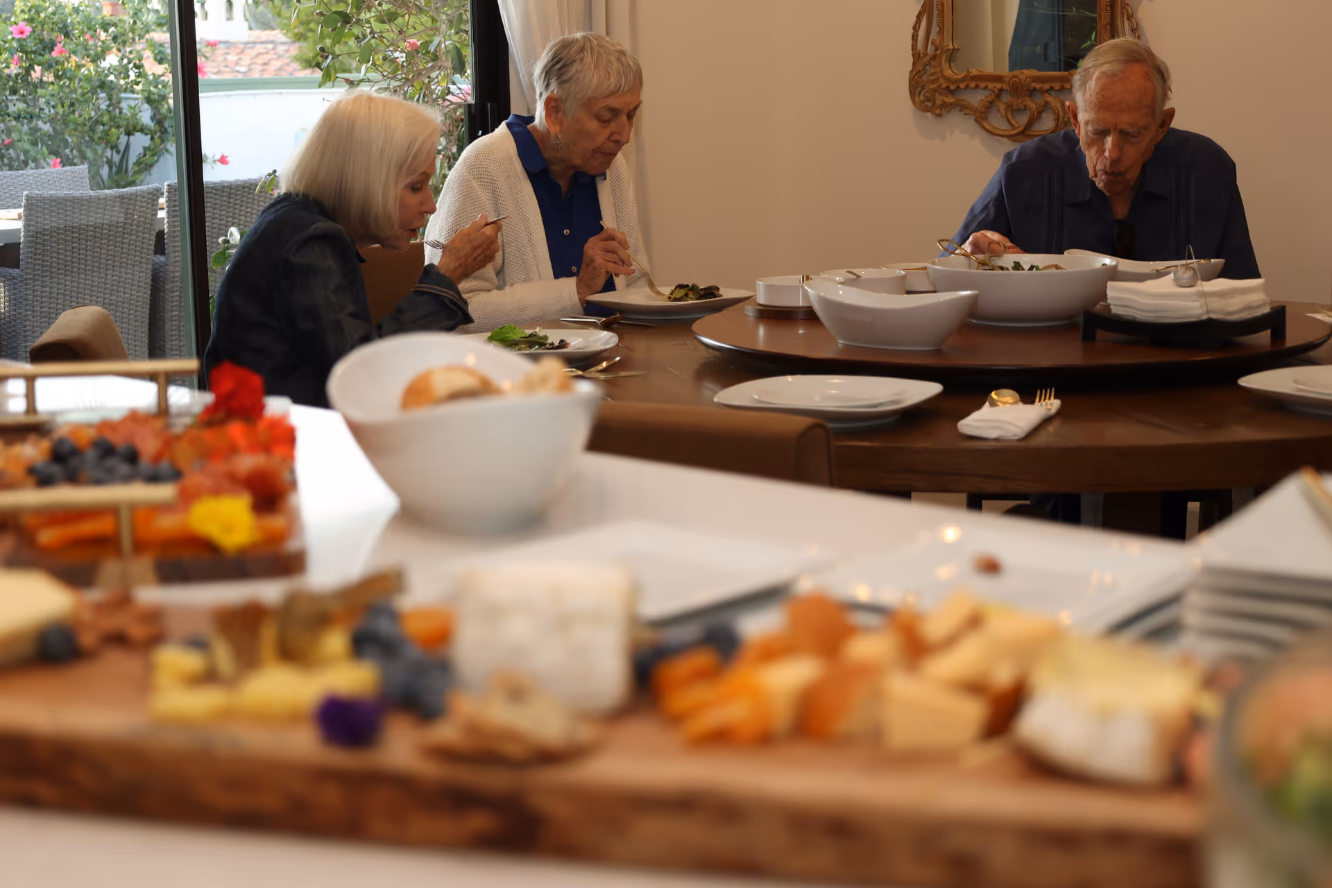 Three seniors dine at a round table with dishes in a bright dining room.