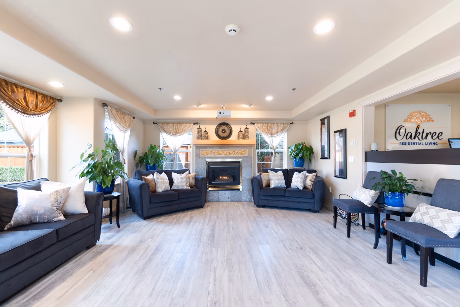 Bright senior living common room with sofas, chairs, plants, and a central fireplace.