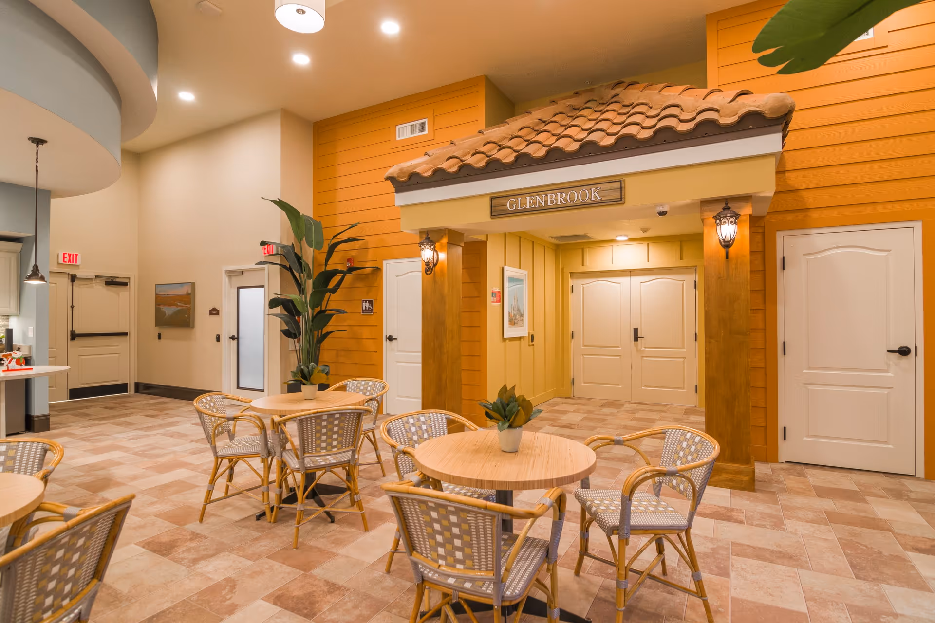A bright and welcoming indoor seating area with round wooden tables and wicker chairs. The walls are painted in warm tones with a decorative structure resembling a small house entrance labeled 'GLENBROOK'. There are plants on the tables and a large potted plant near the wall. The floor is tiled with a beige and light brown pattern, and there are multiple doors and exit signs visible.