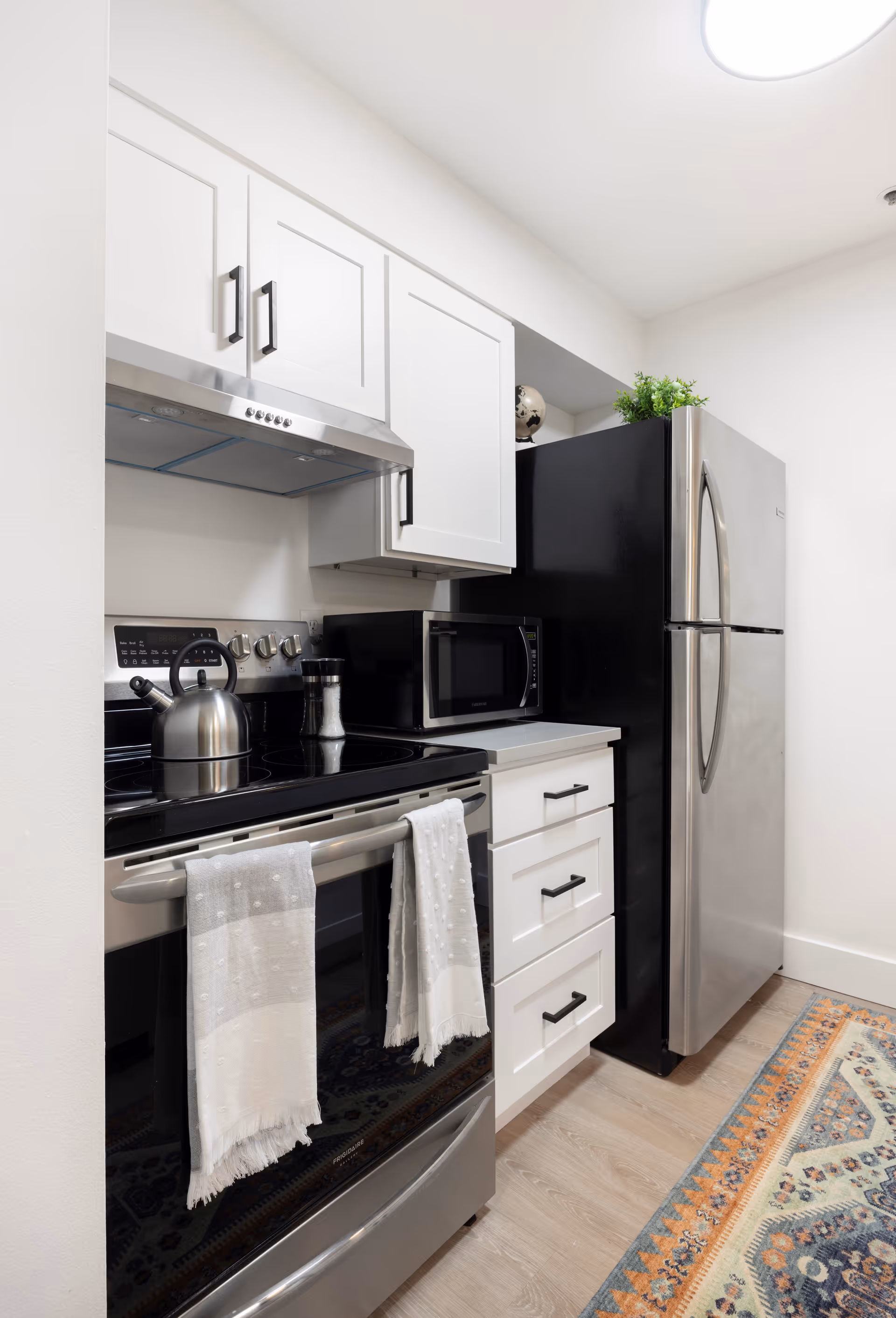 A modern kitchen featuring a stainless steel electric stove with two white towels hanging on the handle, a stainless steel kettle on the stove, a black microwave on the countertop, white cabinets with black handles, a stainless steel refrigerator, and a colorful patterned rug on the wooden floor.