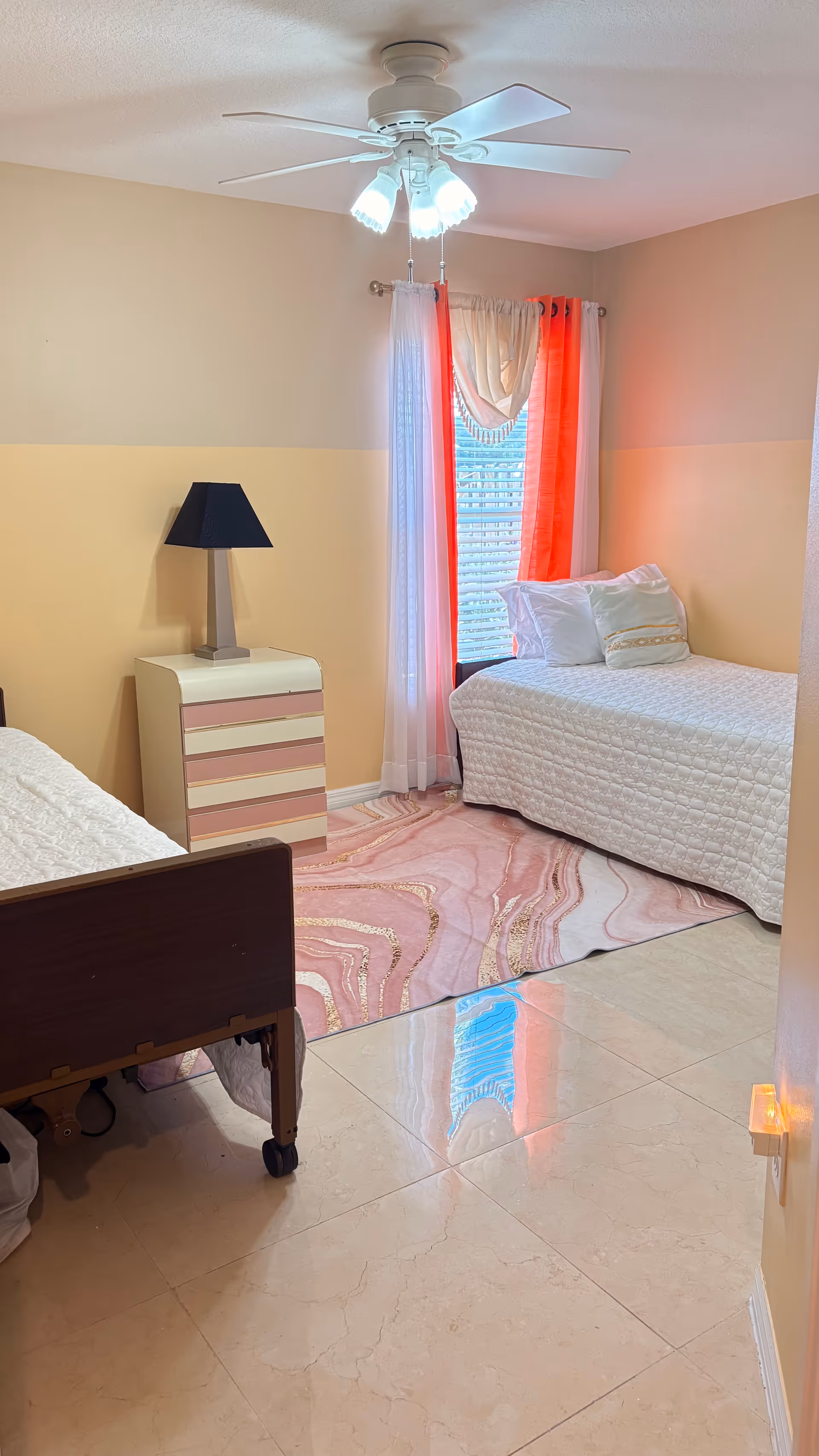 Pastel-toned senior living bedroom with two beds, a nightstand, ceiling fan, and pink rug.