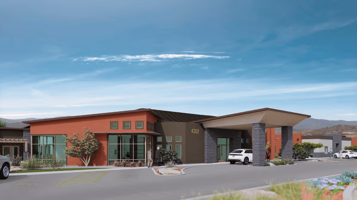 Modern single-story assisted living facility with a covered entrance, parked cars, and desert landscaping under a blue sky.