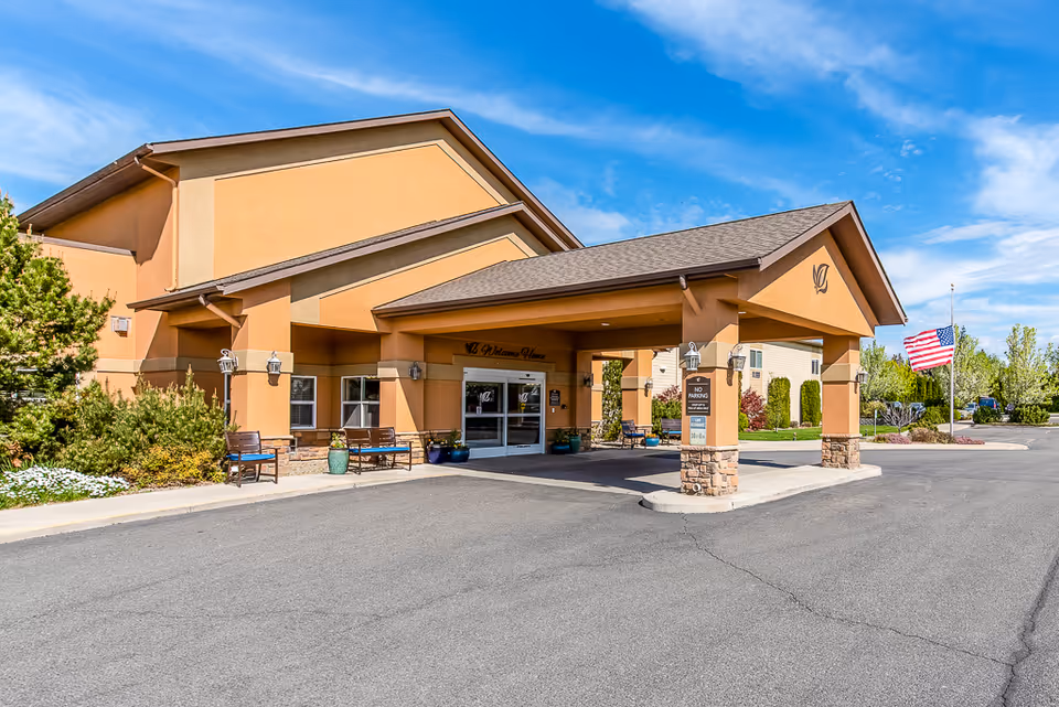 Exterior view of Ellensburg Senior Living facility entrance with a covered drop-off area, benches, potted plants, and an American flag flying on a flagpole under a blue sky with some clouds.