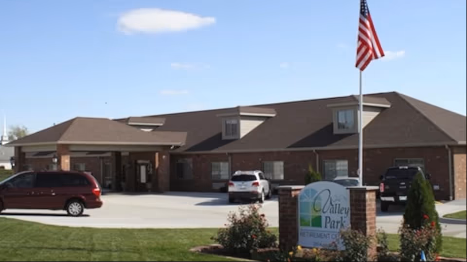 Exterior view of Valley Park Retirement Center building with a driveway, several parked cars, an American flag on a flagpole, and a sign with the facility's name surrounded by landscaping.