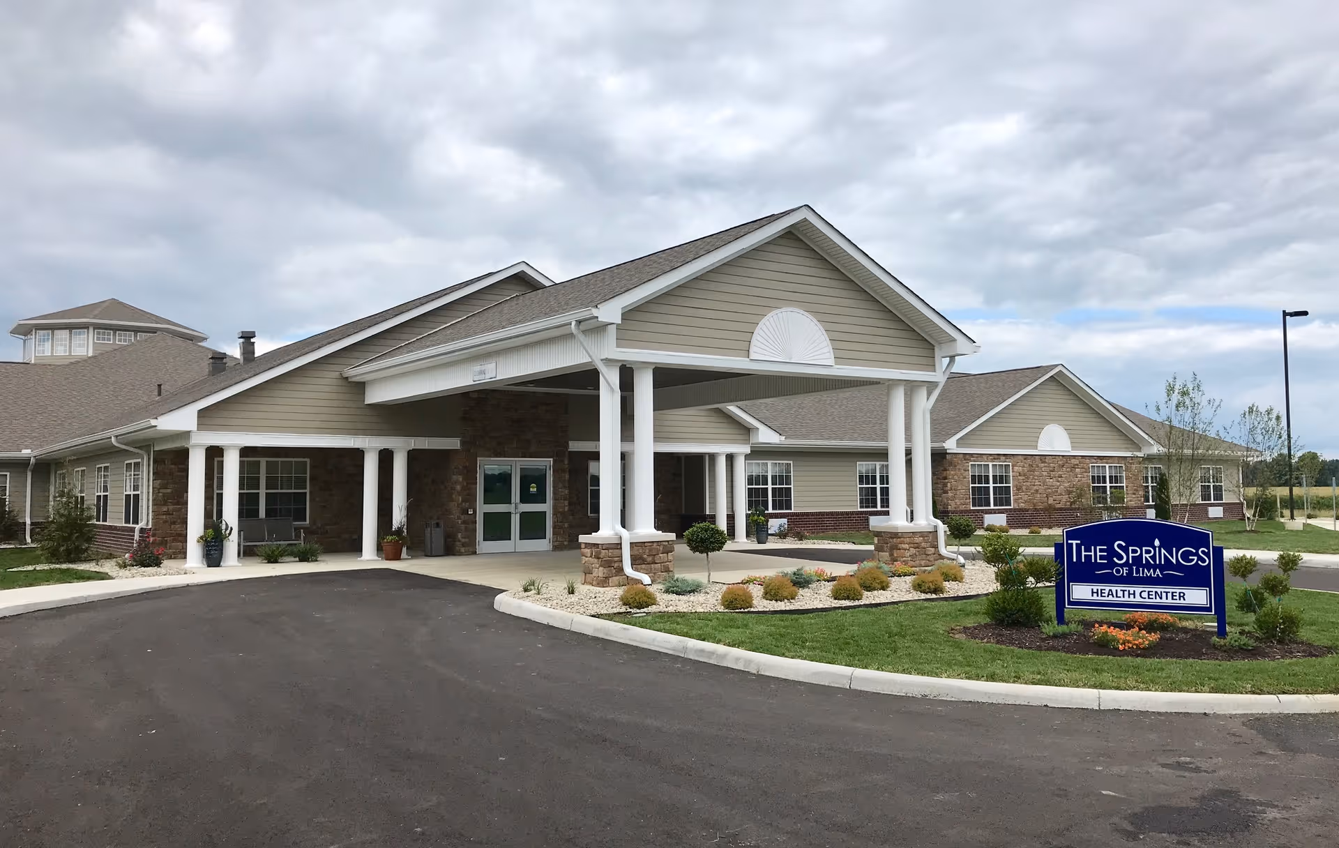 Exterior view of The Springs of Lima Health Center building with a covered entrance, landscaped garden, and a sign displaying the facility's name. The sky is cloudy.