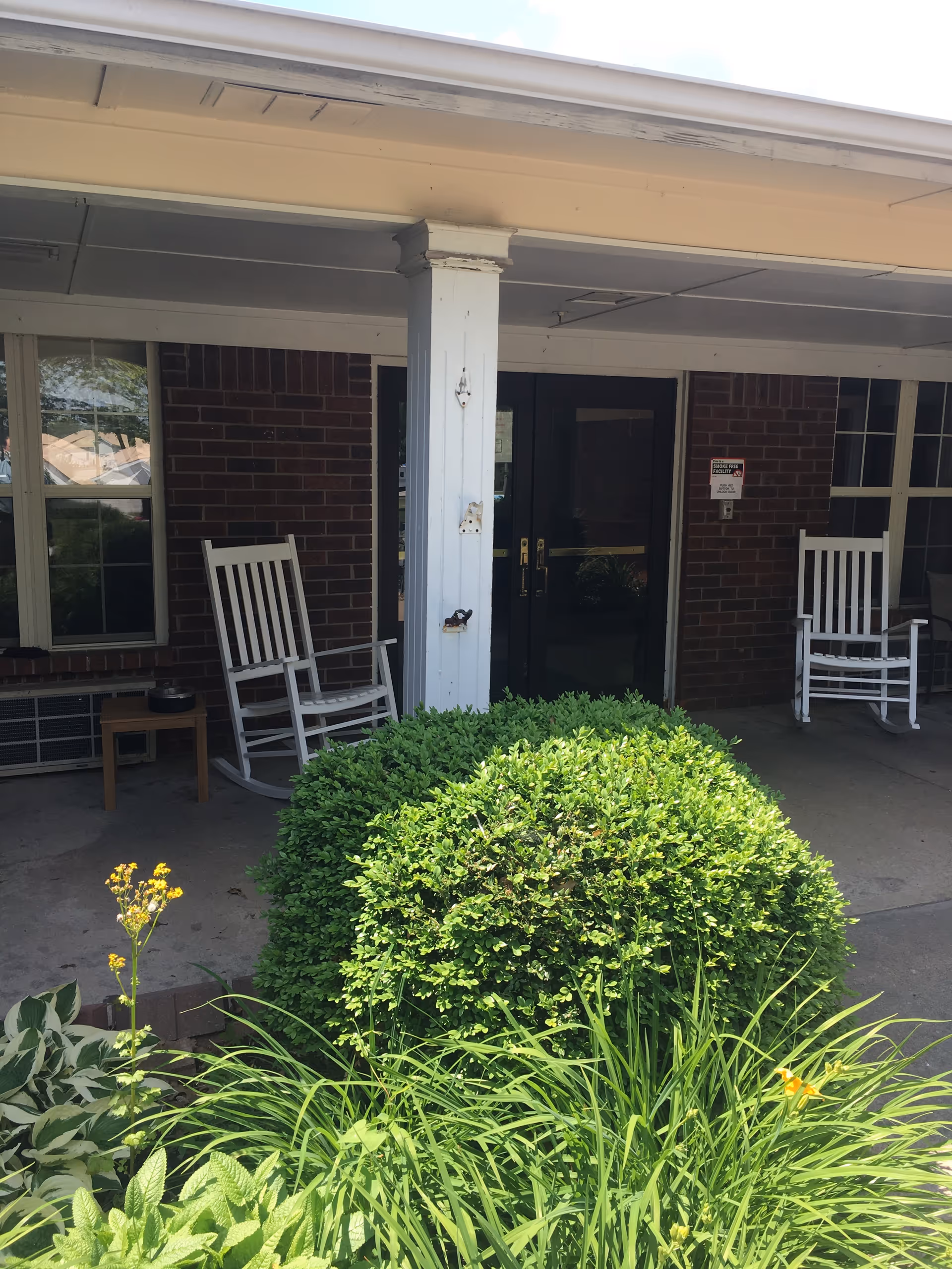Covered brick entrance with double doors, two white rocking chairs on the porch and a trimmed shrub and plants in the foreground.