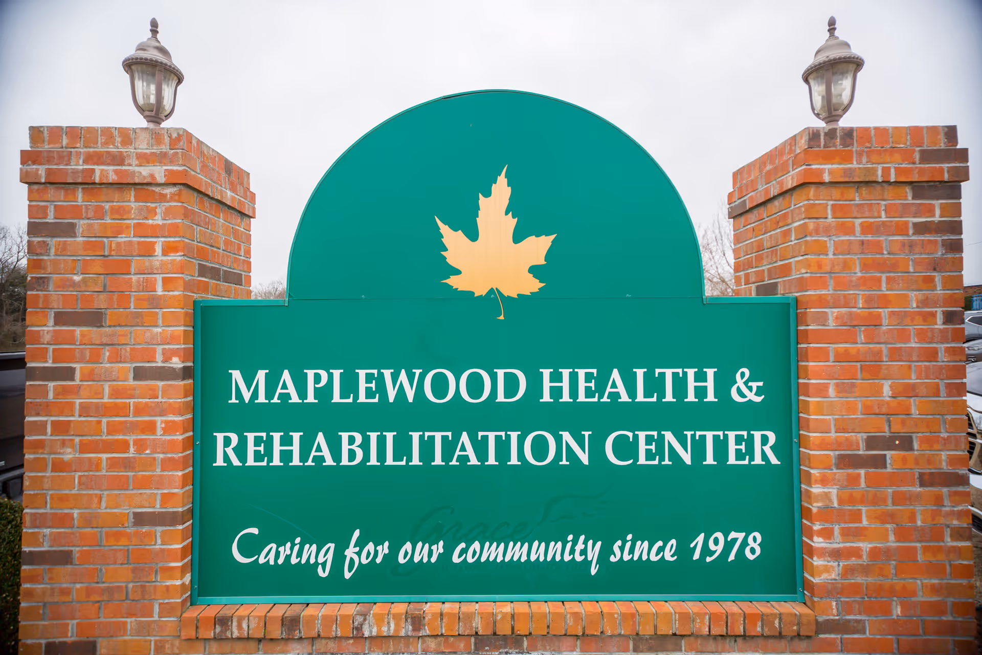 A green sign with a yellow maple leaf at the top, mounted on a brick structure with two lamps on either side. The sign reads 'Maplewood Health & Rehabilitation Center Caring for our community since 1978'.