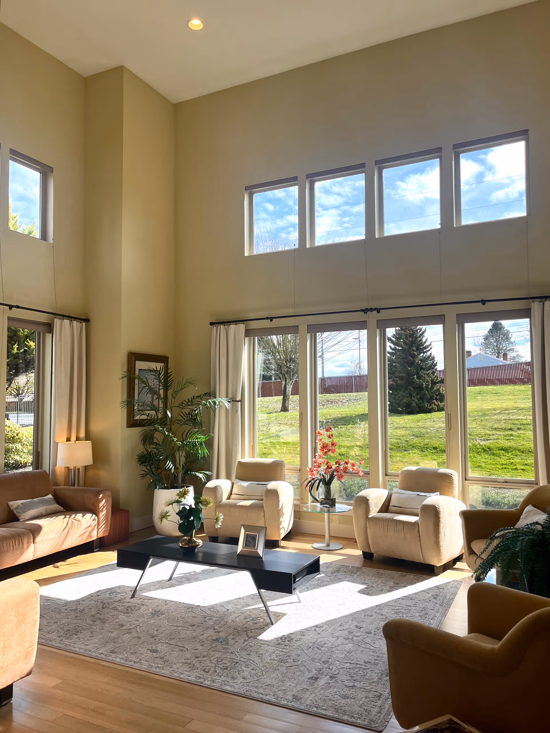 Sunlit living room in a senior facility with cream seating and large windows.