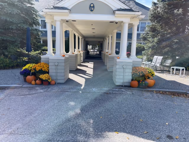 Covered entrance walkway to a building with white columns and a peaked roof displaying the number 78. The walkway is decorated with pumpkins and colorful potted flowers on both sides. There are trees and chairs visible on the right side of the entrance.