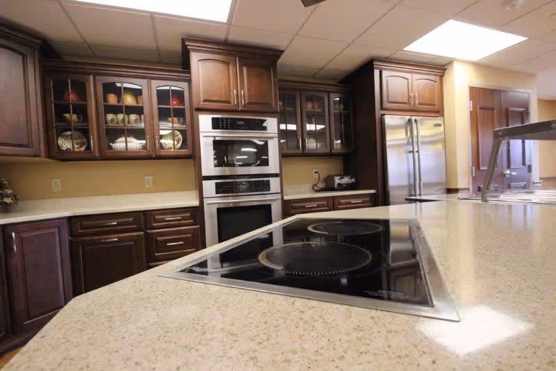 Spacious kitchen with a large island cooktop, stainless steel double ovens and refrigerator, and dark wood cabinets with glass-fronted upper cabinets.