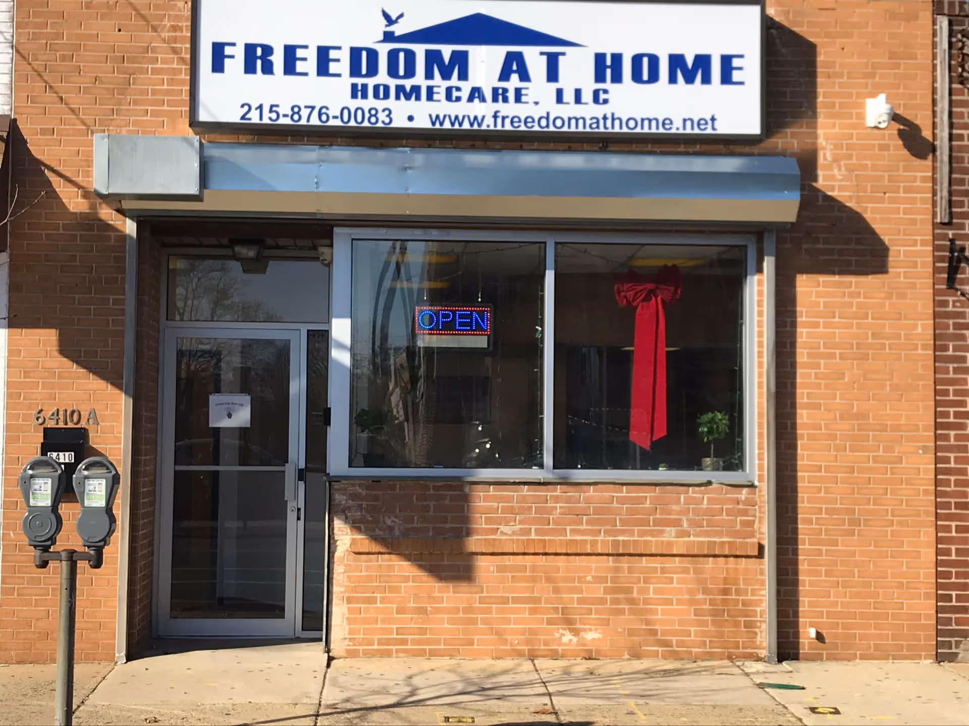 Brick storefront with Freedom At Home HomeCare LLC sign, large window, and OPEN neon sign.