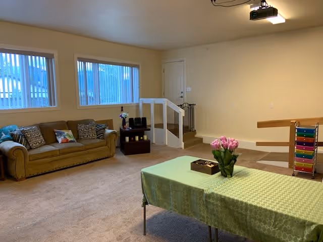 A cozy living room with a beige carpet and cream-colored walls. There is a brown sofa with patterned cushions along the left wall under two windows with vertical blinds. A small dark wooden side table with decorative items is next to the sofa. In the foreground, a rectangular table covered with a green patterned tablecloth holds a vase of pink flowers and a small wooden box. To the right, there is a colorful xylophone toy and a staircase with a white railing leading up to a closed door. A ceiling-mounted projector is visible above.
