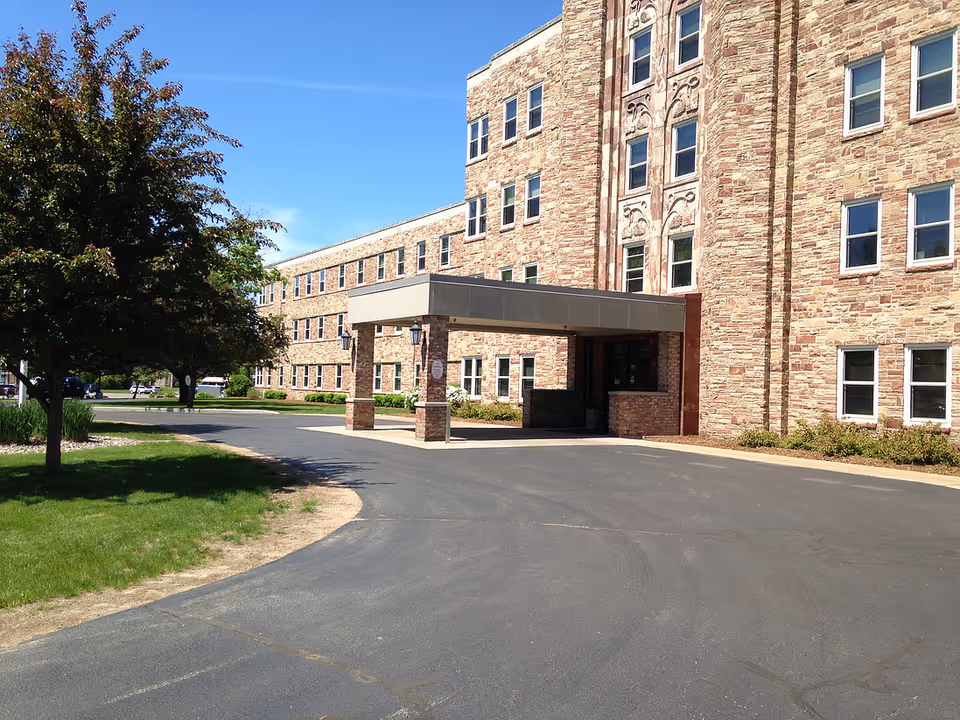 Exterior view of a multi-story assisted living facility with a stone facade and a covered entrance driveway. There are trees and green grass on the left side under a clear blue sky.