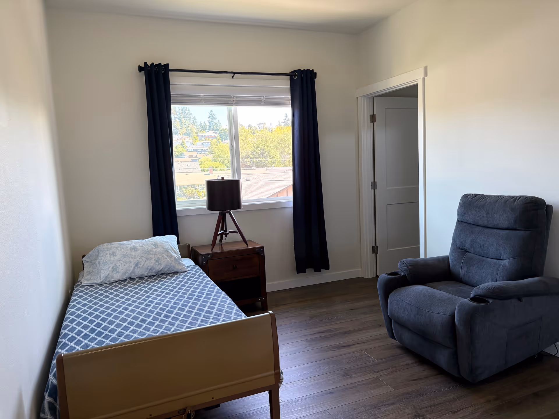 A small furnished bedroom with a single bed, window, nightstand, lamp, and a recliner.