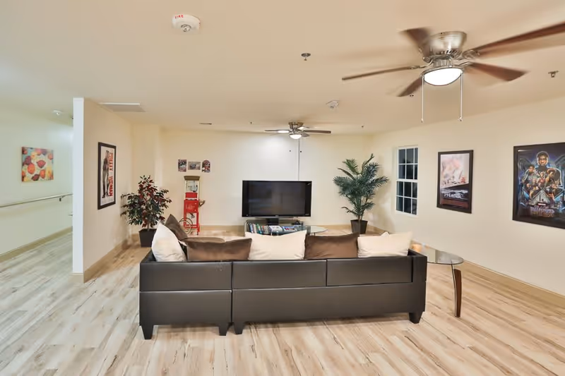 A communal living room with a black sectional sofa facing a wall-mounted TV, ceiling fans, framed posters, and potted plants.