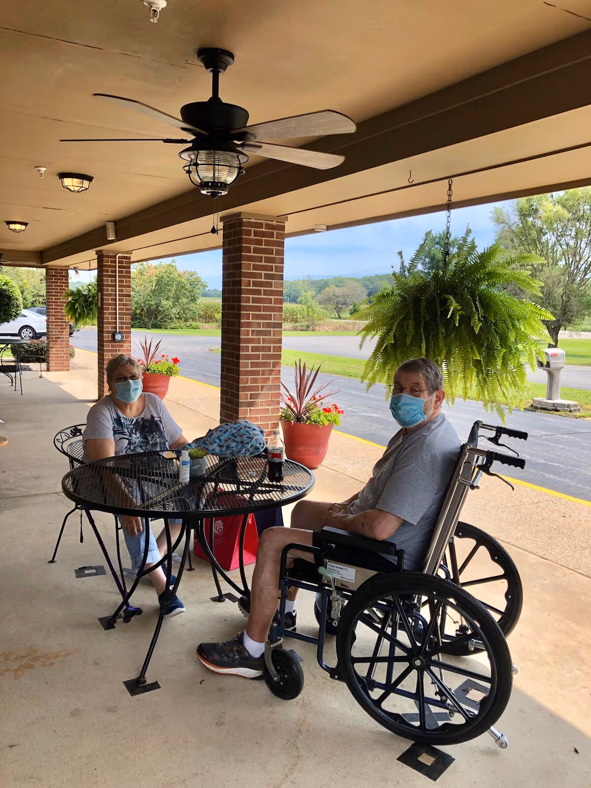 Two elderly individuals wearing face masks sitting at a round metal table on a covered outdoor patio. One person is seated in a wheelchair, and the other is sitting on a metal chair. There are hanging plants and potted plants around, with a view of a parking lot and greenery in the background.