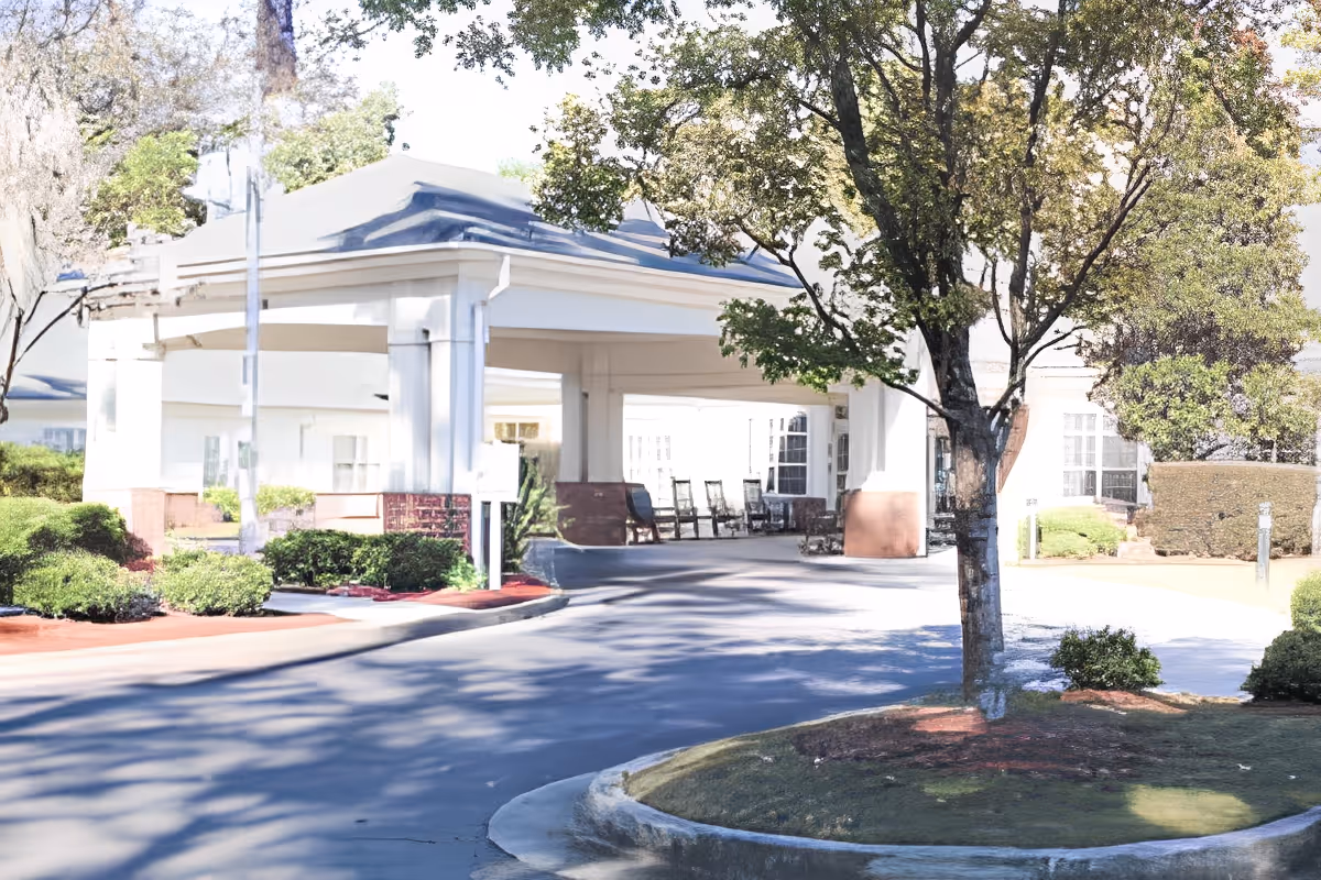 Entrance area of a senior living facility with a covered driveway, several rocking chairs under the covered area, surrounded by trees and landscaped bushes.
