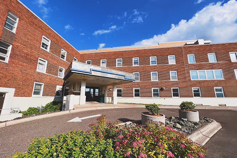 Brick senior living facility exterior with entrance canopy, driveway, and circular planters.
