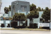 Exterior view of a gray facility building with an arched entrance and trees along a sidewalk.