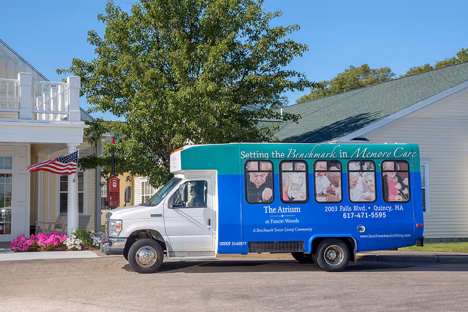 A blue and green shuttle bus for The Atrium at Faxon Woods parked outside the facility entrance with an American flag and flowers.