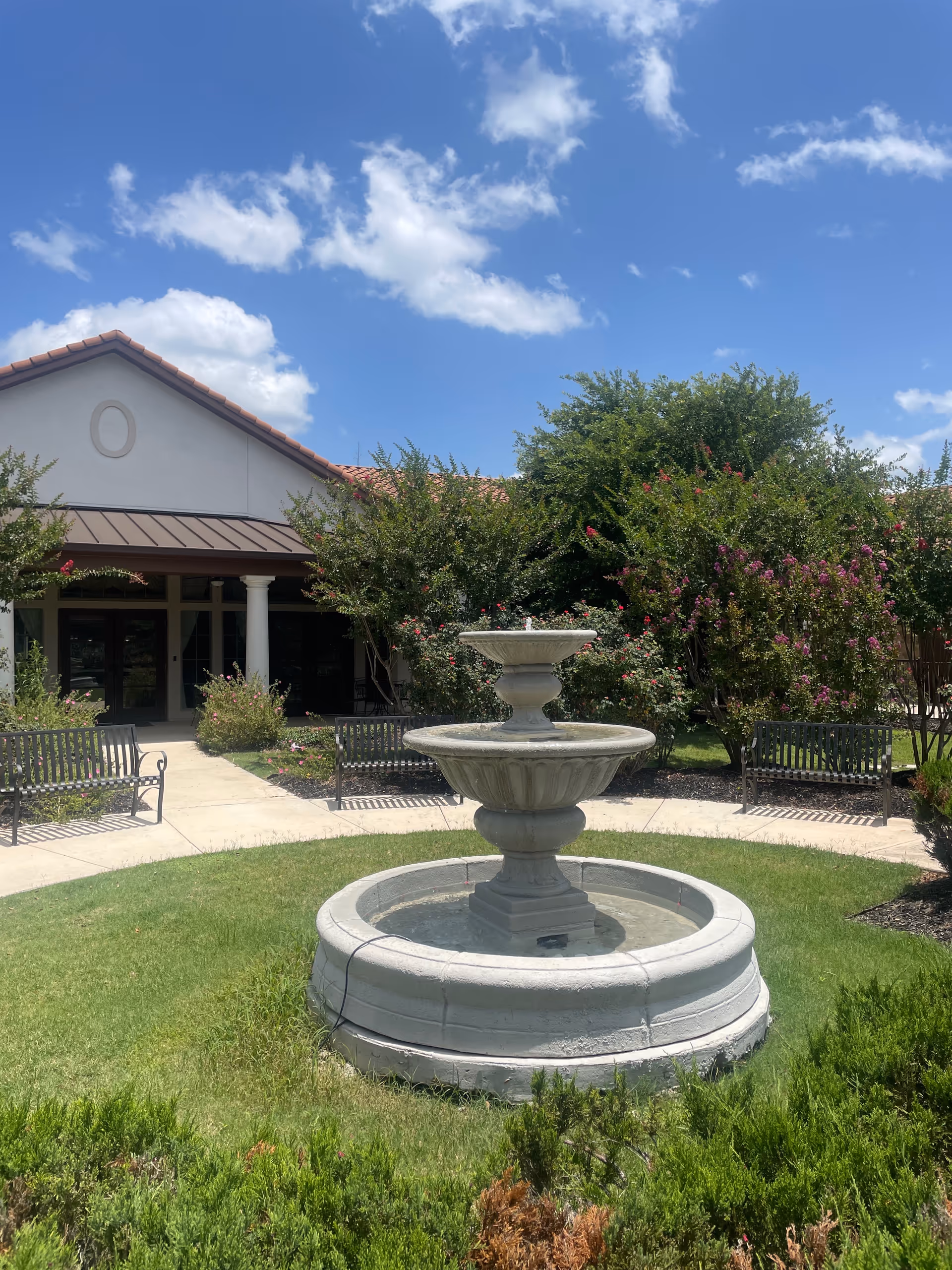Stone fountain in a sunny outdoor courtyard with benches and flowering trees near a building.