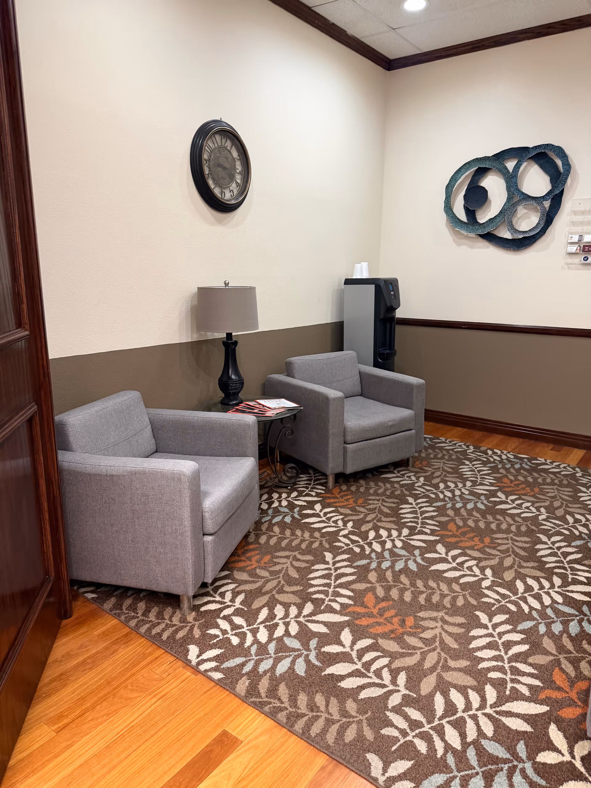Two gray armchairs with a side table, lamp, clock, and water cooler in a waiting area.