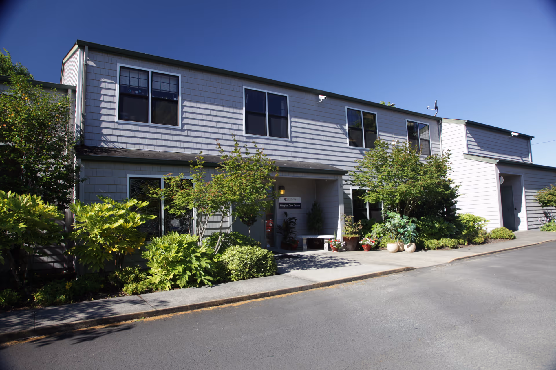 Two-story grey building with shrubs, entrance, and windows along a paved street.