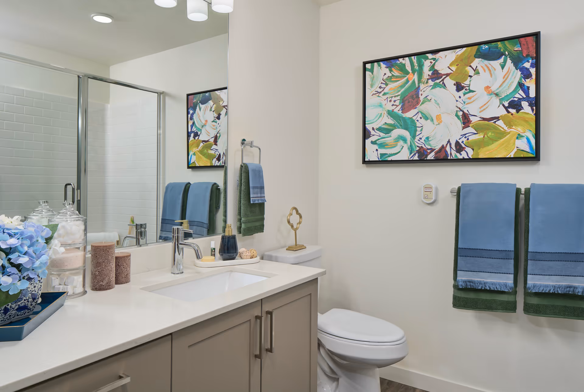 Bright modern bathroom with a sink vanity, mirror, glass shower, toilet, towels, and colorful floral wall art.