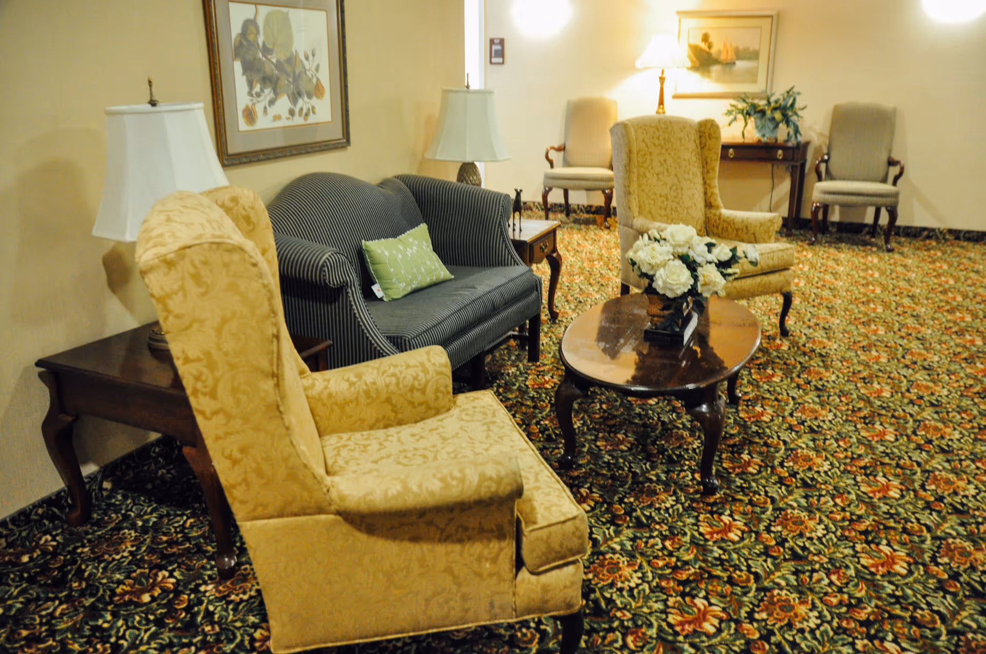 A cozy sitting area in an assisted living facility featuring two yellow patterned armchairs, a dark striped loveseat with a green pillow, a wooden coffee table with a white floral arrangement, two side tables with lamps, and additional chairs against the wall. The room has a floral carpet and framed artwork on the walls.